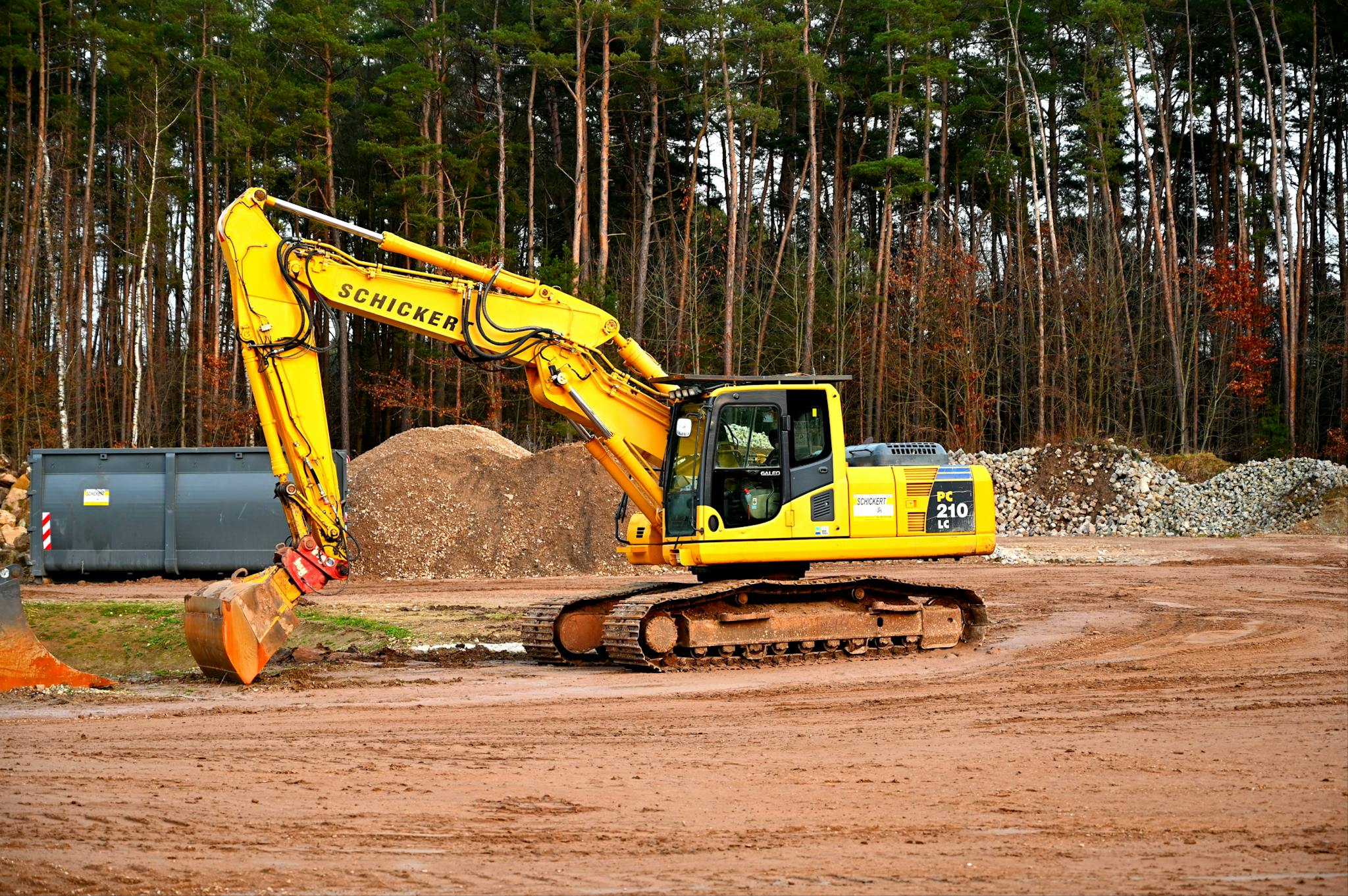 Home A yellow excavator at work in a forested construction site, surrounded by dirt and gravel.