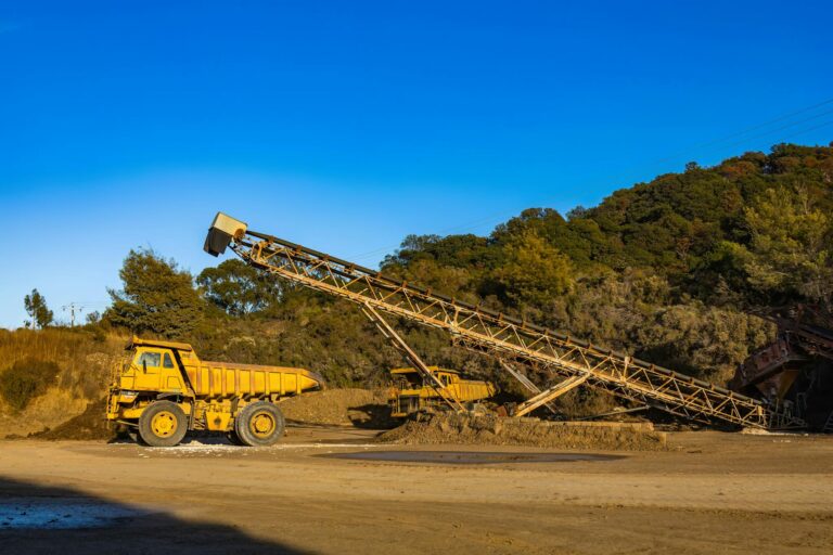 Yellow dump trucks and conveyor machinery in a sunny mining site with clear blue sky.